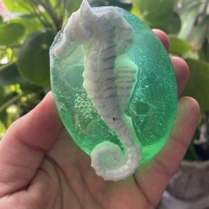 dłon trzymająca zielone, przeźroczyste mydło, z konikiem morskim z przodu. Na tle zielonych liści